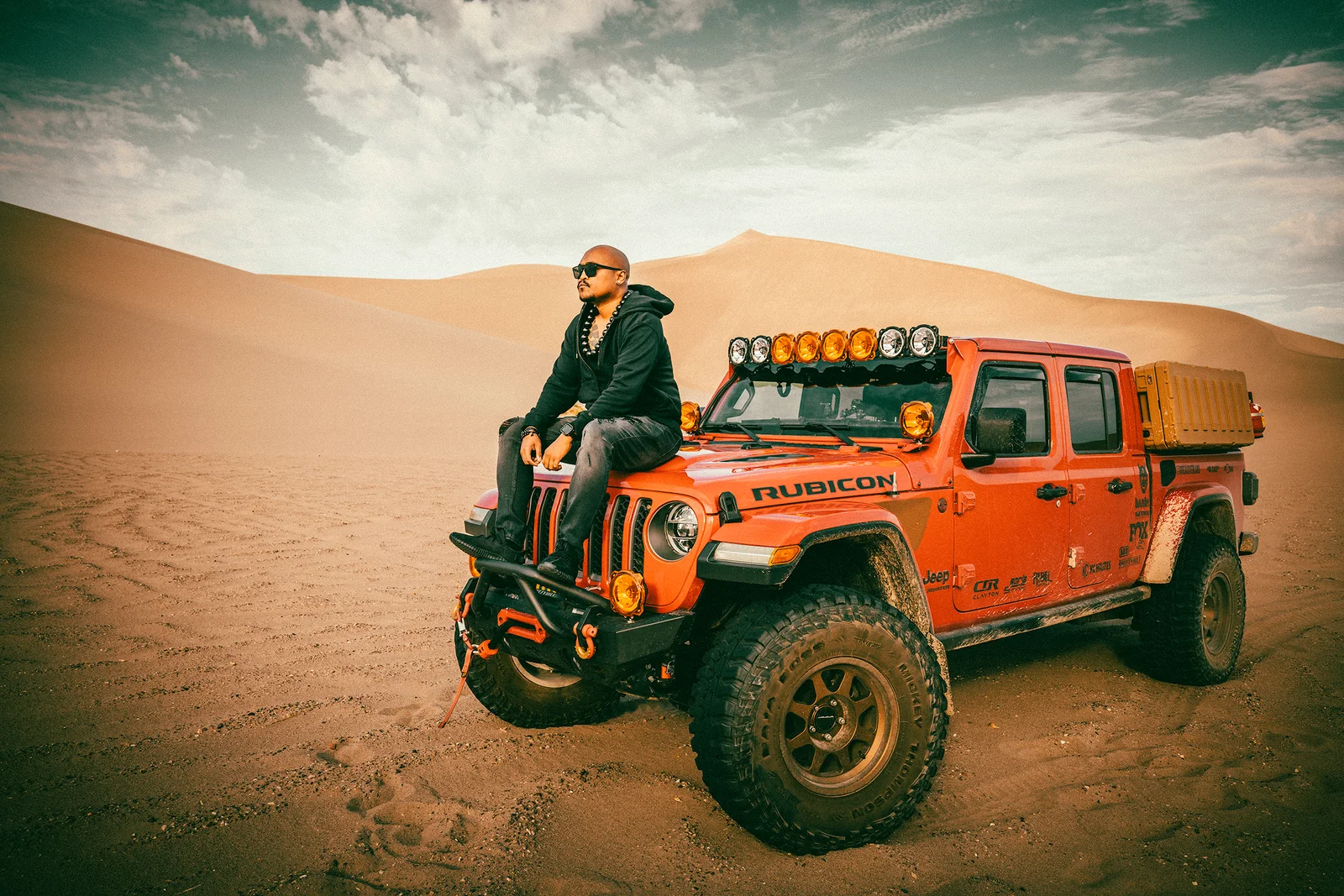 Patrick Breaux seated on the hood of an orange Jeep Gladiator Rubicon with light bar and off-road build, parked on open sand dunes under a wide sky