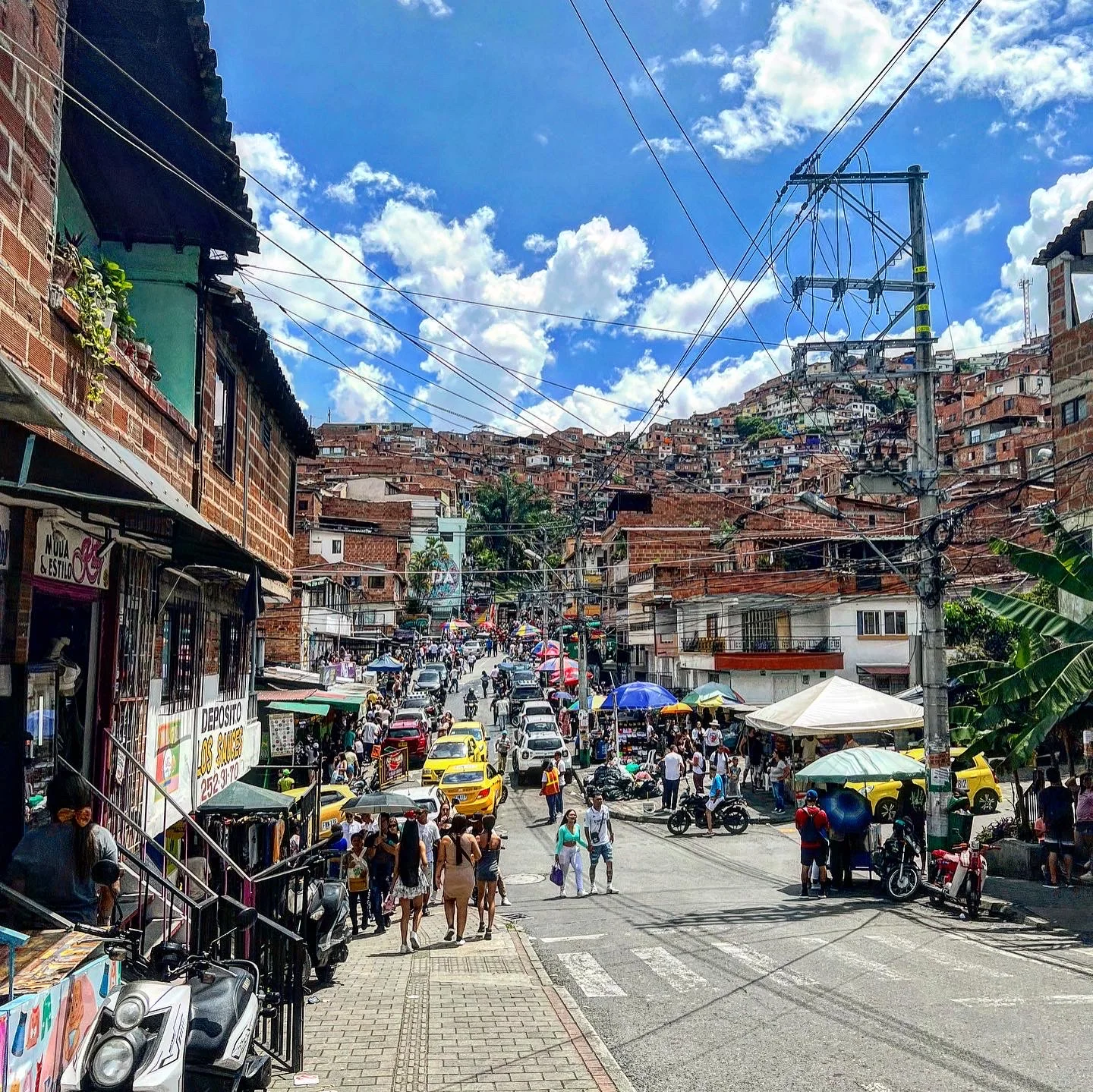 Busy hillside street in a Medellín comuna, brick houses climbing the slope, market stalls and motorbikes under a bright sky