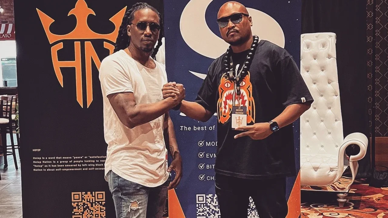 Patrick Breaux and Hotep Jesus shaking hands at HotepCon, HotepNation and Syscoin event banners behind them