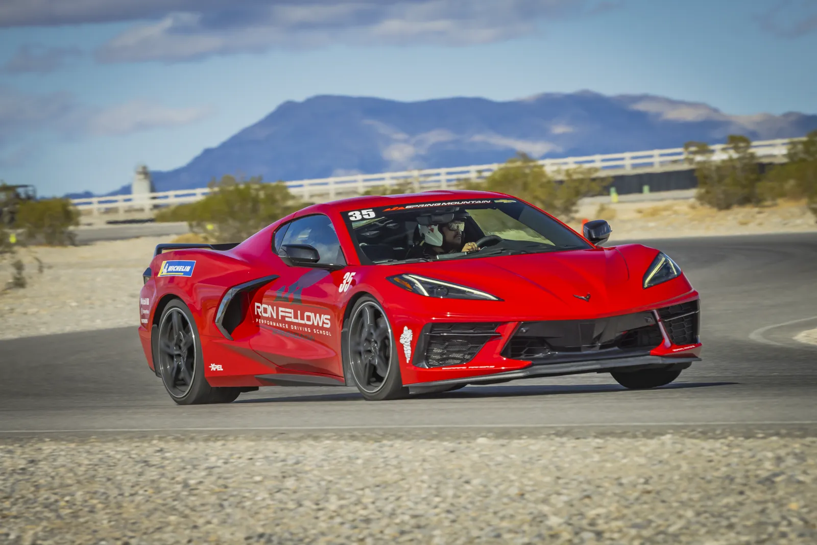Red Chevrolet Corvette C8 mid-corner on Spring Mountain West Track, Ron Fellows door decals visible, Mojave mountains in the background